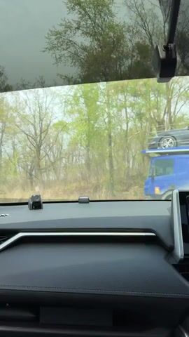 The view from inside a car shows a long line of car carrier trucks stopped on a rural road. The trucks are loaded with various passenger vehicles.  The road is straight and stretches into the distance.  Trees line the side of the road. The sky is partly cloudy. The interior of the car is visible, including the dashboard with a camera mounted on it.  The car is stopped behind the car carrier trucks.