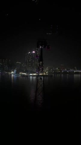 The video shows a nighttime cityscape reflected in water, viewed from a high vantage point, potentially a cable car or elevated structure. In the foreground, a metal support structure of a cable car system is visible, with moving cars. The skyline in the background consists of numerous buildings with lights, indicating a densely populated urban area. The water reflects the lights from the buildings, creating a shimmering effect. The overall impression is one of a modern city at night.