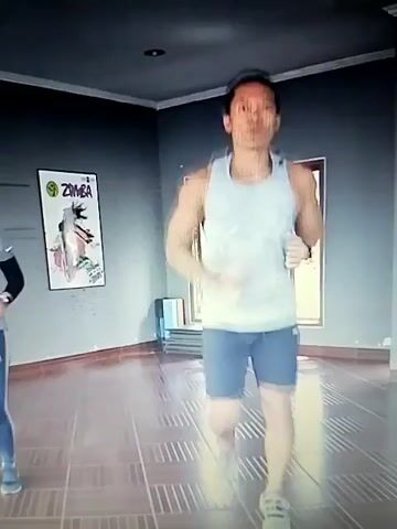 A man in a white tank top and blue shorts is jogging in place in a room. He is facing the camera and appears to be exercising. There is a poster on the wall behind him that says "ZUMBA." The room has tiled floors and gray walls.