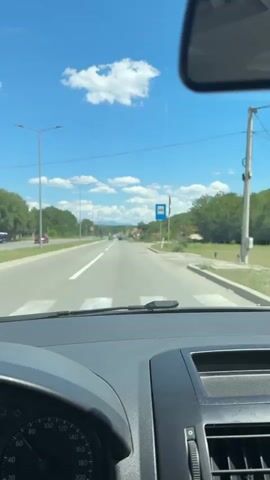 The video is shot from the perspective of someone in a car looking forward. The car is driving on a paved road with white lane markings. There are streetlights and trees lining the road. In the distance, there are more cars and buildings, suggesting a rural or suburban area. The sky is blue with scattered white clouds. There are speed limit signs visible, indicating a speed limit of 60. The interior of the car, including the dashboard and windshield wipers, is visible in the foreground.