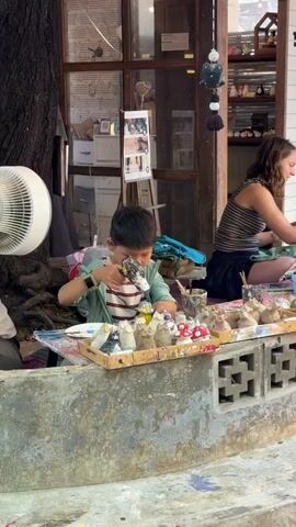 A young boy and a woman are painting figurines outdoors. The boy, wearing a striped shirt and green jacket, is focused on painting a white figurine. He holds a paintbrush in his right hand and a small cup of yellow paint in his left. In front of him are several unpainted and partially painted figurines, including mushrooms and animal shapes, arranged on a tray. A white fan is visible to his left. The woman, with long brown hair, is seated to the right, also engaged in painting. She wears a striped tank top and dark skirt. Behind them is the entrance to a shop with shelves displaying various items. The scene appears to be a creative workshop or market stall.