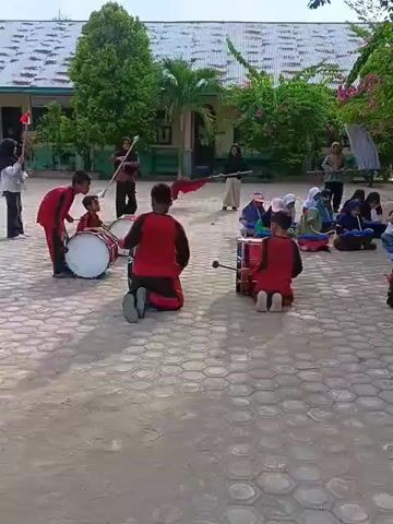 A group of children are practicing in a courtyard. They are wearing red and black uniforms. Some of the children are playing drums, while others are sitting and listening. There are also some adults in the background, observing the practice. The courtyard is paved with hexagonal tiles, and there are trees and buildings around. The children are all young, and they appear to be engaged in their practice.