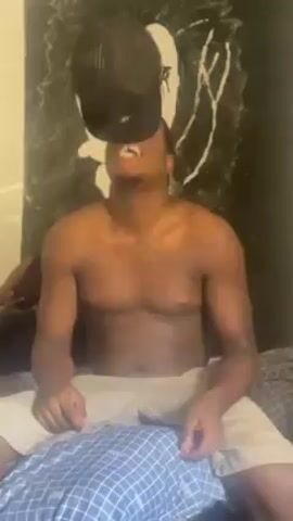 A shirtless Black man is sitting on a bed, holding a black baseball cap. He smiles, puts the cap in his mouth, and pulls it out. Behind him, a woman sits holding a phone. There is a painting of a man with long hair on the wall behind them. The man is wearing light-colored shorts. The bed has a blue and white patterned cover.