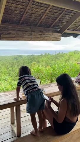 Here is a description of the video. A woman with long dark hair sits on a wooden bench, focused on her phone.  Next to her, a small child wearing a striped shirt and blue shorts leans on a wooden table, gazing out at the view. The view includes lush, green trees and a distant ocean, framed by a thatched roof above. The scene appears to be taken on a porch or raised platform.