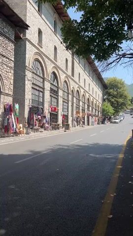 A street scene shows a long, multi-story stone building with arched windows lining one side. Small shops or stalls are set up in front of some of the ground-floor arches, displaying textiles and other goods. A paved road runs alongside the building, with a yellow line marking the edge of the lane. Several cars are visible on the road, with some moving and others parked. Trees with green leaves hang over the scene from the right side, and a hill is visible in the distance. The sky is clear and blue.