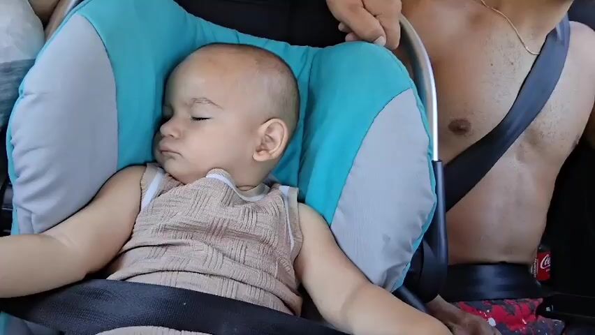 A baby is sleeping in a car seat. The baby is wearing a light brown tank top. A person's hand is visible holding the car seat. To the right of the baby, a shirtless person with a seatbelt is visible. A bracelet with the name "Kemal" is visible on the wrist of a person. The environment appears to be inside a vehicle.