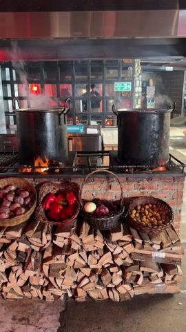 The video shows a cooking setup with two large, blackened pots steaming on a brick platform over a gas-fired cooking range. Below the cooking surface, neatly stacked firewood is arranged. Four woven baskets sit on the firewood, filled with different vegetables: red onions, red bell peppers, a mix of white and purple onions, and small potatoes. Above the pots, there is a stainless steel ventilation hood. The backdrop includes mirrored panels reflecting a person moving behind the scenes, possibly a chef. An exit sign is visible in the background. The scene is well-lit, with a warm, inviting feel.