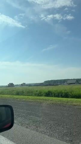 The video shows a view from a moving vehicle. The foreground is a grey asphalt road with a white line on the left side. A black rearview mirror is visible on the lower left side of the frame, with a hint of red interior. The middle ground features a vibrant green field of grass or crops. In the background, several houses with red and brown roofs are scattered across the landscape. A line of trees forms a dark green border behind the houses. The sky is a clear blue with scattered white clouds. The sun is visible in the upper left corner, casting a bright light. The overall scene depicts a rural landscape on a sunny day.