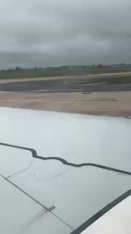 The view from an airplane window shows a white wing with black seam lines and rivets. Outside, the ground is a patchwork of gray concrete runway, green grass, and a hint of distant buildings under an overcast sky. The airplane appears to be moving along the runway. The scene is stationary, filmed from inside the aircraft.