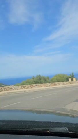 A car drives down a road next to a stone wall with trees and houses on one side and the ocean on the other. The sky is blue with white clouds. A second car is parked on the side of the road. The car continues to drive down the road, passing more houses and trees. A tall tree is on the right side of the road.