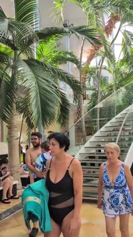 The video shows an indoor tropical atrium with palm trees and lush vegetation. A winding staircase with glass railings is a prominent feature. Several people are visible, including two women in swimsuits in the foreground. One woman, on the left, wears a black one-piece swimsuit with mesh inserts and carries a blue towel. The other woman, on the right, is in a blue and white patterned one-piece swimsuit. Further in the background, other individuals can be seen walking and standing on a balcony area, and a woman in a sheer cover-up walks down the stairs. The overall atmosphere appears to be a resort or spa environment with a focus on natural elements.