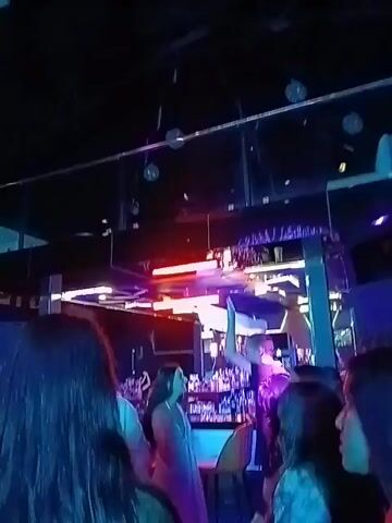 The video shows a crowded nightclub with people dancing under flashing multicolored lights.  The camera moves around, capturing glimpses of the crowd and the club’s interior, including a bar and a raised platform. The mood appears festive and energetic. Some individuals are clearly visible, while others are blurred due to movement and focus. The overall atmosphere suggests a lively night at a club.