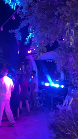 The video captures an outdoor nighttime event with a crowd of people dancing under purple and blue lights. There is a stage with lighting equipment and possibly a DJ booth visible on the right. The atmosphere appears festive, with guests dressed casually and engaging in dancing. The setting seems to be a courtyard or an open-air venue, with trees and possibly some architectural elements in the background.