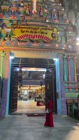 The video showcases a vibrantly colored Hindu temple situated within a cave. The temple entrance is adorned with intricate carvings and colorful decorations. A woman wearing a red dress walks into the temple. The camera pans up to show the high ceiling of the cave. The ceiling is composed of natural rock formations with light and dark coloration.