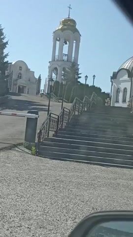 A security camera captures a view of an entryway to a religious complex. A white barrier gate is partially lowered, blocking a paved road. To the right of the road, a grey gravel pathway leads to a wide, grey, stone staircase with black metal railings. The stairs ascend towards a white bell tower with a golden dome topped with a cross. Behind the bell tower, a larger white building with a golden dome is partially visible. Further to the left, another white building with a domed roof is situated on a higher elevation. The sky is clear and blue, and the scene is bathed in bright sunlight. Lush green trees and manicured bushes line the pathways and surround the buildings. The camera appears to be positioned low to the ground, possibly from within a vehicle, as a dark, curved object, likely a rearview mirror, is visible in the bottom right corner of the frame. There are no people visible in the video.