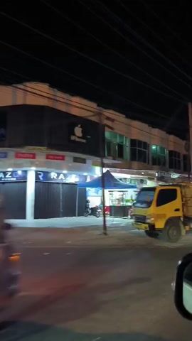 The video pans across a street scene at night. On the left, a yellow dump truck is parked. Behind it, a building with a white facade has several storefronts. One storefront has an Apple logo and the word "iPhone" above it. Another has the word "RAZER" in large letters. A blue canopy is set up in front of one of the stores. Further down the street, more storefronts are visible, including one with the word "COFFEE". There are also food carts and motorcycles parked along the street. The building across the street also appears to have multiple levels and windows. The overall impression is a commercial area at night.