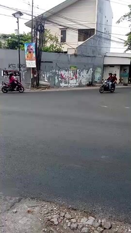 The video shows a street scene with several motorcycles passing by. The road is paved and appears to be an intersection. On the left side of the road, there is a building with a gray facade and some posters on the wall. There are also utility poles and wires. The sky is overcast. The motorcycles are all moving in different directions, with some carrying one or two riders. The riders are wearing casual clothing, and some are wearing helmets. The overall impression is of a busy street in a city.