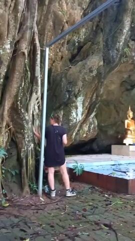 Here is a description of the video:
The camera pans to the right to show a golden Buddha statue placed on a raised platform inside a cave. In the foreground, a woman in a black dress stands beside a large tree and a metal frame.  The platform is decorated with blue tiles. The ground around the platform is covered with leaves and moss.  The cave walls are rocky and textured. Beyond the cave entrance, there is a dense forest. The lighting is somewhat dim inside the cave, but brightens as the camera pans to show the forest.