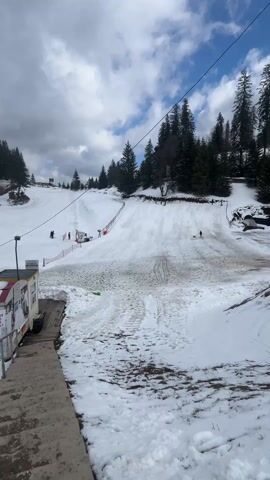The video presents a daytime view of a snow-covered ski slope in a mountain environment. From an elevated vantage point looking down a set of concrete steps, a small building is visible to the left of the frame with signage written on it. The main focus is the snowy slope, which has several sets of tracks indicating previous skiers. A lift station is situated partway up the slope with a red safety fence around it, and an employee dressed in a red uniform can be seen standing by the station. Several people are seen skiing on the slope. The background includes pine trees and a cloudy sky. The overall atmosphere is typical of a ski resort during the day.