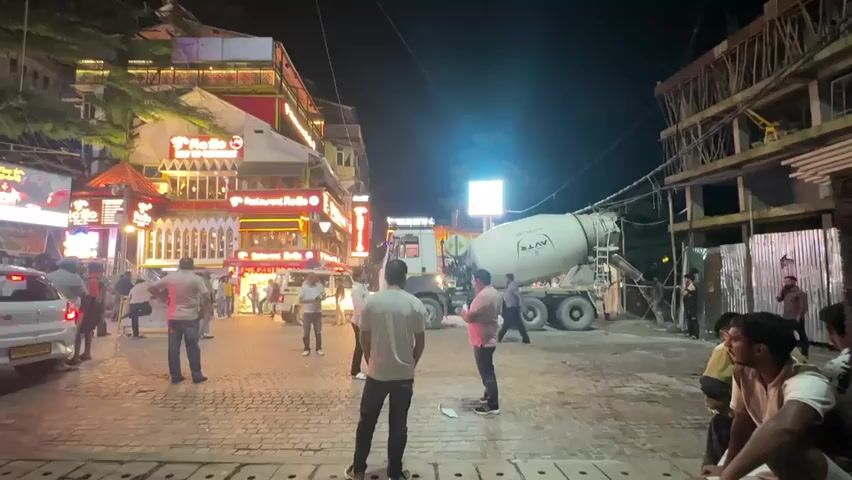 The video captures a street scene at night, illuminated by artificial lights from buildings and construction sites. A cement mixer truck is parked in the middle of a paved street, with its drum tilted as if in operation. Several people are visible around the truck and on the sidewalks. On the left side of the street, there are multi-story buildings with glowing signs, suggesting they are shops or restaurants. Some individuals are standing or walking, while others are gathered in groups. To the right, a construction site is evident with scaffolding and building materials. The overall atmosphere appears to be one of activity and ongoing work despite the late hour.