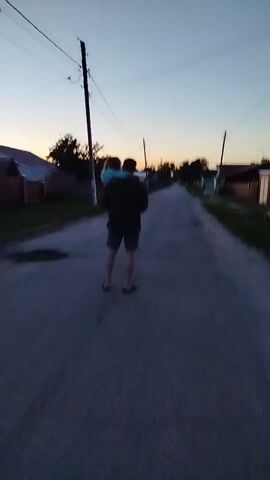 A man walks down a road carrying a young child in his arms as the sun begins to set. The man is wearing a dark jacket, shorts, and flip-flops. The child is wearing a blue hooded sweatshirt. The road appears to be in a rural or suburban area with houses on either side. Power lines and poles are visible along the road. The sky is a gradient of orange, pink, and blue.