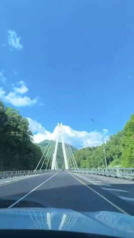A point-of-view shot from inside a vehicle shows a modern cable-stayed bridge under a bright blue sky with scattered clouds. The road is asphalt with white and yellow lane markings. The bridge has two tall, white, A-frame pylons, with numerous cables extending to the road deck. The bridge is surrounded by lush green trees and mountains. The camera is positioned on the dashboard of the vehicle, with the windshield and part of the car's interior visible in the foreground. The video appears to be captured while driving across the bridge.