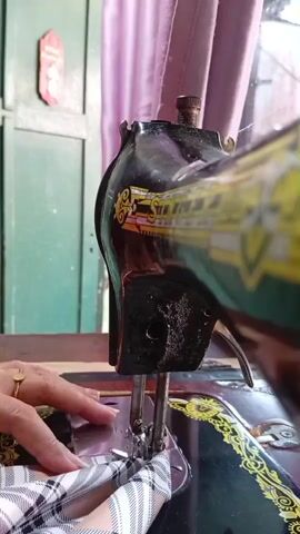 A close-up shot shows a person's hands operating a vintage sewing machine. The machine is black with gold detailing, and a plaid fabric is being fed under the needle. The background is slightly blurred, with a purple curtain and a greenish wall visible.