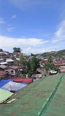 The video shows a view of a dense residential area with many houses clustered together on a hillside. The houses have a variety of roofing materials, including corrugated metal in colors such as green, red, and blue, and some appear to be made of concrete or wood. Lush green trees are interspersed among the buildings, indicating a tropical or subtropical climate. The sky is a clear, bright blue with scattered white clouds. The overall impression is of a vibrant community situated in a hilly landscape under a sunny sky.