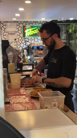 A man is preparing food in a restaurant. The man has a beard and is wearing glasses and a black t-shirt. He is wearing gloves and working on a plate of food, spreading red sauce with his fingers. Behind him is a deli slicer, a coffee machine, and a lit-up sign that says "Totoca". The countertop is covered in a red and white checkered paper. There is a plastic container with ingredients on the counter as well. The background includes a black and white geometric wallpaper.