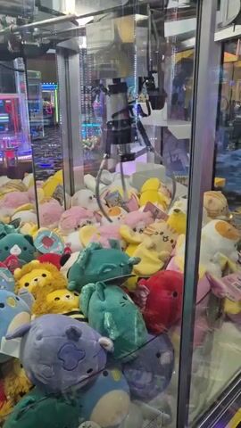 A claw machine filled with plush toys. The claw is moving down towards the toys. In the background, other arcade games are visible.