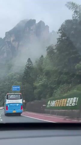 The camera, positioned within a vehicle, captures a road winding through a mountainous, foggy landscape. A blue and white bus travels ahead, its taillights visible. The road is bordered by lush green trees and a grey guardrail. In the distance, towering rock formations are partially obscured by mist. Chinese-style red lanterns hang from lamp posts along the road. The sky is overcast, contributing to the hazy atmosphere.