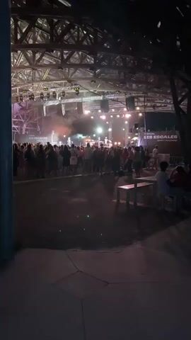 The video captures an outdoor music concert at night. A large crowd of people is gathered in front of a brightly lit stage under a large, exposed wooden roof structure. Stage lights illuminate the stage and cast a warm glow over the audience. Smoke or haze is visible on the stage, adding to the atmosphere. Banners with the text "LES ESCALES" are visible on the right side of the stage. In the foreground, there are empty benches and a paved area. The overall scene suggests a lively and energetic event.
