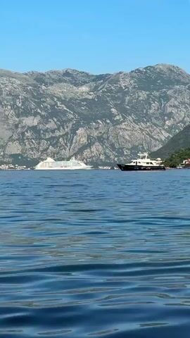 The video shows a wide shot of a body of water with a large white cruise ship and a smaller dark-hulled yacht in the distance. Mountains covered in greenery and rock formations form the backdrop. The water in the foreground is calm with ripples reflecting the sunlight. The cruise ship is positioned to the left, while the yacht is closer to the center right. The sky is clear and blue. The camera pans slightly to the right.