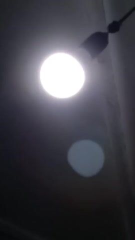 The video shows a light bulb illuminating a room. The light source is bright and round, and a slightly dimmer circular reflection is visible below. The bulb appears to be attached to a fixture on the ceiling. The room itself is dark, with the ceiling surface partially visible. An audio track is present, but the content is not described.