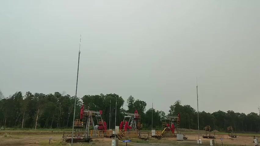 The video shows an outdoor scene with multiple oil pumpjacks in operation. The sky is overcast and gray. In the foreground, there is a dirt or gravel area with some equipment and signage. In the middle ground, there are at least four red and yellow oil pumpjacks, some of which appear to be actively pumping. Tall metal poles with what look like antennas or weather instruments are visible near some of the pumpjacks. In the background, there is a dense line of trees. The overall environment suggests an oil field or industrial area.