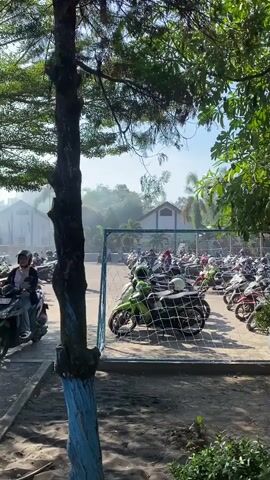 The video shows a parking lot filled with many motorcycles under a bright, sunny sky. A tall tree with green leaves is in the foreground, obscuring part of the view. Beyond the motorcycles, there's a soccer goal with a net, and behind that, several buildings and lush green trees. Some people are visible walking among the motorcycles, and a few are riding them. A small amount of smoke is visible in the distance, possibly from a vehicle or a controlled fire. The ground appears to be a mix of sand and pavement, with some scattered debris.