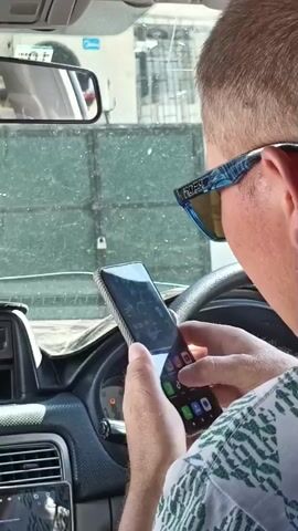 A man wearing sunglasses is seen from the side, looking down at his smartphone. He is in the driver's seat of a car. The dashboard and steering wheel are visible. The man is holding the phone in his right hand and appears to be scrolling or interacting with it. The phone has a case with a textured pattern. The screen shows app icons, including one that looks like TikTok. 

The setting appears to be daytime, with light filtering into the car. Outside the car windshield, a dusty, green metal gate with a padlock is visible. The overall impression is that the car is parked or stopped. The man's face is partially obscured by his sunglasses and the angle, but he seems focused on his phone. The scene is likely in Kazarman, Kyrgyzstan, given the location context. There is no overt indication of emotion, but the focus on the phone suggests a moment of personal engagement or distraction.