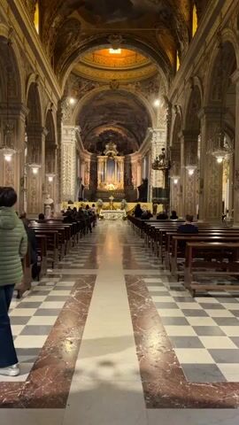 The video shows the interior of a church. It's an ornate building with a checkered tile floor and rows of wooden pews. People are seated in the pews, facing the altar. The altar is decorated with gold trim, candles, and a statue. Above the altar is a mural painted on the ceiling. Someone is speaking in the background.