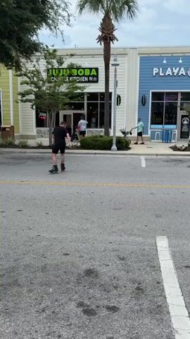 A person is rollerblading down a street in front of several storefronts. The person is wearing a black t-shirt and black shorts and is moving from left to right in the frame. To the left of the rollerblader, there is a storefront labeled "JUJU BOBA CHINESE KITCHEN". To the right of that, there is a storefront labeled "PLAYA BOWLS". Further to the right, there is a storefront labeled "The Eye Gallery". A palm tree is visible behind the "JUJU BOBA" storefront, and other trees and greenery are scattered throughout the scene. The sky is overcast.