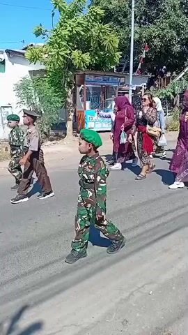 A group of children in camouflage uniforms and green berets are marching on a paved road under a clear blue sky. The children appear to be in elementary school. Behind them, a group of women are walking, some wearing headscarves and traditional clothing. The surroundings include houses with white and green walls, trees, and a street vendor stall. The atmosphere appears festive, possibly a parade or a school event.