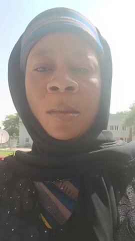 A woman wearing a black hijab and a patterned headscarf is centered in the frame, looking directly at the camera. She has a nose piercing. Behind her, there is a building with white and beige walls, and a few figures wearing uniforms are walking. The video appears to be taken outdoors in daylight. The woman's expression is neutral, then she slightly smiles.