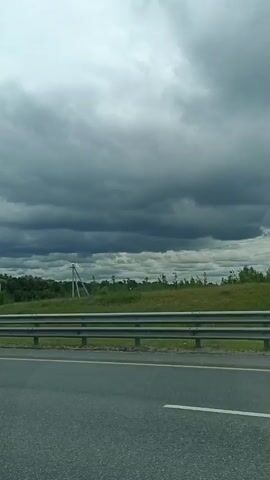 This is a vertical video clip from a moving vehicle, likely a car, traveling on a highway. The scene depicts a rural or semi-rural landscape under a cloudy, overcast sky. 

The primary subjects are the road and the surrounding environment. In the foreground, a white sedan is driving on the highway. The road surface is asphalt with white lane markings. A metal guardrail separates the highway lanes from the shoulder. Beyond the guardrail, there is a grassy area with trees and bushes. In the distance, there are more trees forming a dense treeline, suggesting a forest or wooded area. A few utility poles with wires are visible interspersed among the trees.

The weather appears to be cloudy, with dramatic, dark grey storm clouds filling a significant portion of the sky. This suggests a potentially stormy or at least unsettled weather condition. The lighting is diffused due to the cloud cover, making it difficult to determine the exact time of day, but it appears to be daytime.

There are two pieces of visible text. On the left side of the frame, a green road sign is mounted on a metal structure. The sign has white text and an arrow pointing right. The visible text reads "ОВ" and "КОВО", suggesting it is providing directions to a location. On the right side of the frame, a black and white billboard or sign is partially visible in the distance. The text on this sign is too small to be clearly read but appears to be a advertisement.

The overall mood conveyed by the video is one of travel through a landscape under a somewhat somber sky. The activity is simply driving, with the camera capturing the passing scenery. The location is suggested to be Новосёлки, Russia, based on external context. No people are directly visible, only the car in which the video is being recorded.