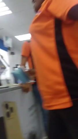 The video shows a brightly lit commercial interior.  Several individuals are present, including two women wearing orange polo shirts with black accents.  The women are standing near a counter with the text "Reno 15 Series" printed on it. The scene appears to be a retail store, with displays of electronics and cell phones visible in the background. A ceiling fan can be seen. The camera moves and focuses on one of the women.