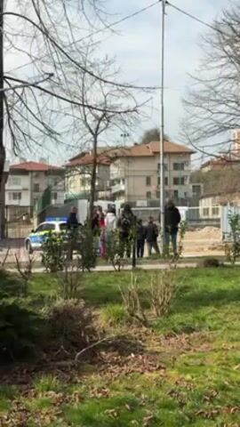 The video shows a group of people gathered near a construction site in what appears to be a park or public space. A police car is parked nearby, and a red truck drives past. A group of people in green uniforms and white head coverings can be seen walking on the sidewalk next to the construction site. The scene is filmed from a distance, with trees and bushes in the foreground. There is sound in the video; possibly music from wind instruments or a trumpet. The weather seems mild, with a mix of sunshine and clouds.
