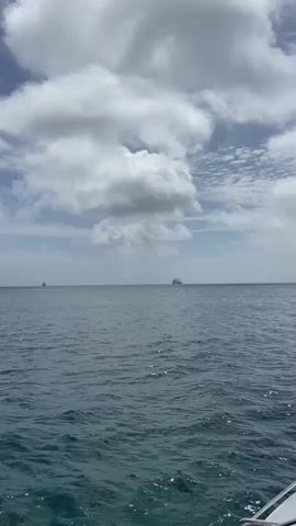 The video begins with a wide shot of the ocean under a cloudy sky. The water is a deep blue-green with gentle waves. In the distance, several large ships are visible on the horizon. As the video progresses, the camera pans slightly to the left, revealing a mountainous landmass in the far distance, partially obscured by clouds. The ships remain visible on the horizon. Closer to the camera, a white catamaran with a tall mast sails across the frame from left to right. The sky remains cloudy with patches of blue. The video ends with the catamaran continuing to move away.