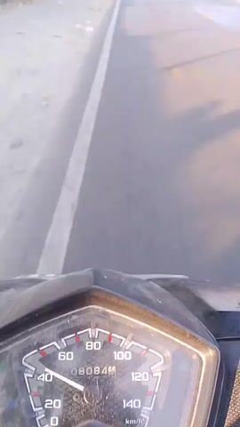 The video begins with a close-up of a motorcycle's speedometer. The needle indicates a speed of approximately 30 km/h. The road ahead is visible, with a white line dividing the lanes. The surroundings appear to be a rural or suburban area with some vegetation and buildings visible in the distance. The video then continues with a first-person perspective from the motorcycle, showing the road ahead and the speedometer. The speed appears to increase slightly as the video progresses, reaching around 40 km/h. Several other vehicles, including cars and motorcycles, are seen on the road in the distance. The sun is shining, casting shadows on the road, suggesting it is daytime. The video concludes with a shot of the road and speedometer, with the speed appearing to hover around 35-40 km/h.