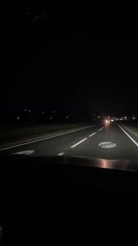 The video begins with a view from inside a vehicle at night, looking out through the windshield. The road ahead is illuminated by the vehicle's headlights and other vehicles in the distance. There are white lines marking the lanes on the asphalt road. On the left side of the road, there is a dark, flat landscape with scattered lights in the far distance. On the right side, the landscape also appears dark and flat, with some lights visible. A phone screen, showing what appears to be a navigation app and possibly a music app, is visible in the lower left corner of the frame. The vehicle is moving forward on the highway. The sky is completely dark. The camera perspective is static, suggesting it is mounted or held steady.