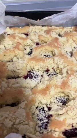 The video shows a close-up of a freshly baked crumb cake with dark red berries visible beneath the crumb topping. The cake is in a baking pan lined with parchment paper. The crumb topping is golden brown and appears to be made of small, irregular pieces of dough. The berries are deep red and appear juicy. The lighting is bright, highlighting the texture and colors of the cake.
