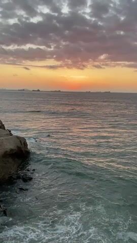 The video shows a view of the ocean at sunset from a rocky cliff. The sky is filled with clouds tinged with pink and orange from the setting sun. The sun itself is a bright orange orb just above the horizon. Several ships are visible in the distance, appearing as dark silhouettes against the horizon. The waves are crashing against the rocks at the base of the cliff, creating white foam. The water is a deep blue-green. The overall atmosphere is peaceful and serene.