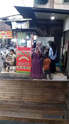 A group of people are gathered around a food stall. The stall has a red and green sign that says SABENA. There are pictures of food items on the sign, including what appears to be fried chicken. A man is sitting on a stool to the left of the stall, holding a notebook and pen. Several women and a child are standing in front of the stall, seemingly ordering food. The environment appears to be an outdoor market or street. There are motorcycles parked in the background. The time stamp indicates the video is at the beginning of the recording.
